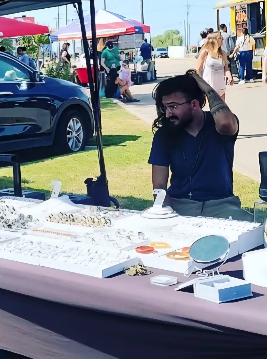 Man at a jewelry booth at an outdoor market with people and cars in the background.