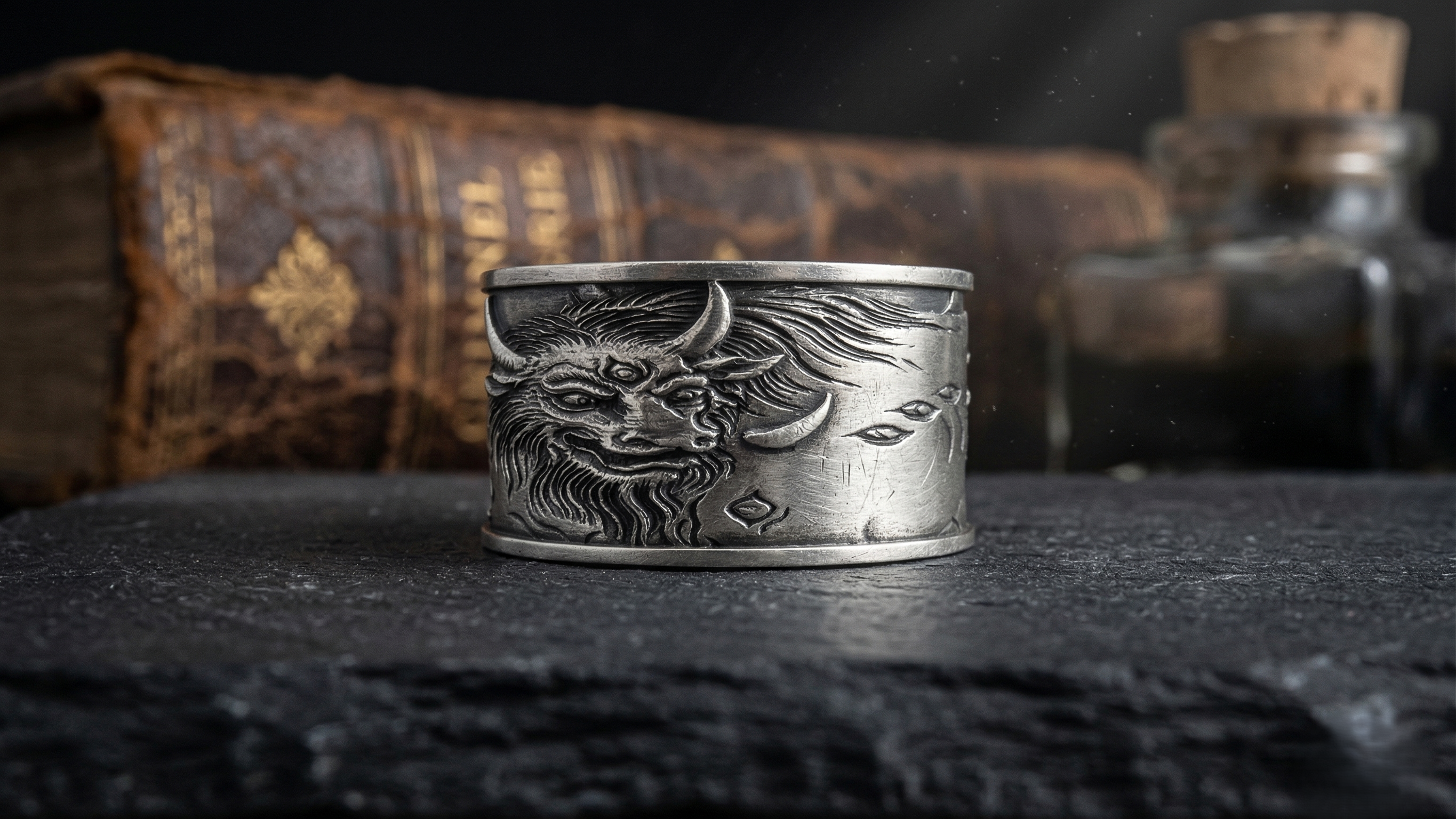 Silver ring with a detailed design on a dark surface with books and bottles in the background